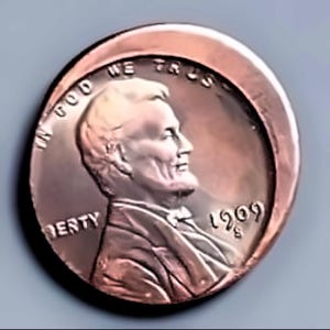 Puede incluir: Una moneda de centavo Lincoln de 1909, de color cobre. El anverso presenta un perfil de Abraham Lincoln, con las inscripciones "IN GOD WE TRUST" y "LIBERTY". El año "1909" es visible. La moneda tiene una forma ligeramente curvada.