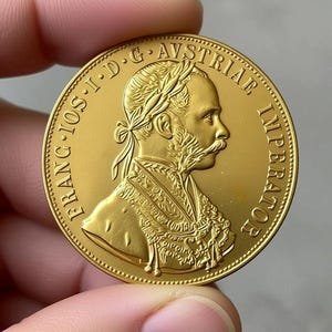 Puede incluir: Una moneda de oro con el perfil de un hombre con una corona de laurel y bigote. La moneda tiene texto alrededor del borde, incluyendo "FRANC IOS I D G AVSTRIAE IMPERATOR". La moneda está sujeta entre dos dedos.