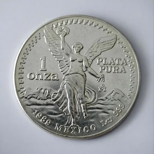 Puede incluir: Moneda de plata con una figura alada sosteniendo una corona de laurel, con las palabras "1 onza" y "Plata Pura". La moneda es de 1989 y está marcada "Mexico".