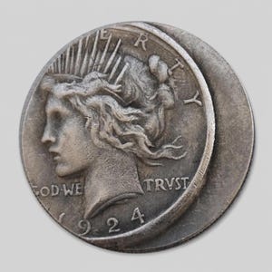 Può includere: Una moneta Peace dollar color argento che presenta il profilo di Lady Liberty con una corona radiante. La moneta mostra il testo LIBERTY, IN GOD WE TRUST e l'anno 1924. La superficie presenta una texture metallica usurata con dettagli in rilievo visibili.