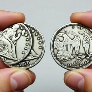 Puede incluir: Dos monedas de plata se sostienen en las manos. Una moneda presenta una mujer sentada con un martillo y el año 1870. La otra moneda muestra un águila con las palabras "OF AMERICA" y "DOL". Estas monedas son probablemente objetos de colección antiguos.