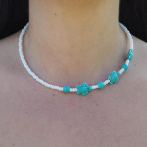 May include: White beaded necklace with turquoise turtle and bead accents. The necklace features three turtle charms and turquoise beads interspersed along the white beaded strand. A simple, elegant design.