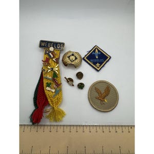 May include: A collection of scouting memorabilia including a Webelos pin bar with yellow, red, and green braided cords, a gold-tone neckerchief slide, a diamond-shaped blue patch, three small metal pins, and a circular tan patch with an eagle emblem.