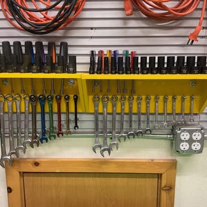 May include: A yellow tool organizer mounted on a wall, holding various wrenches, sockets, and screwdrivers. The tools are neatly arranged, with a mix of sizes and colors. Electrical cords and outlets are also visible.