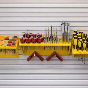 Könnte beinhalten: Eine Sammlung von Werkzeugen, die auf gelben Regalen angeordnet sind. Zu den Werkzeugen gehören Bohrer, Schraubendreher und andere Hardware. Die Regale sind an einer Wellblechwand befestigt. Es gibt auch orangefarbene Schleifpads und rote Winkel.