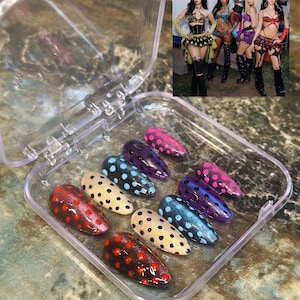 May include: A set of ten almond-shaped press-on nails in a clear plastic case. Each nail features a distinct color base, including red, beige, blue, and purple, all decorated with contrasting polka dots. An inset photo shows five people.
