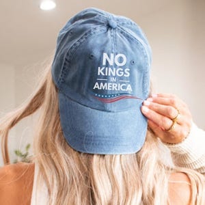 May include: A blue baseball cap with the text "NO KINGS IN AMERICA" in white. The cap has a red and white stripe design below the text. The cap is worn by a person with long blonde hair.