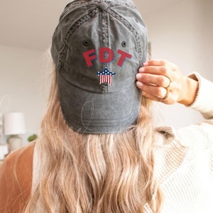 May include: A gray baseball cap with the letters "FDT" in red and a star with red and blue stripes. The cap is worn by a person with long blonde hair.