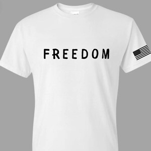 May include: White t-shirt with the word "FREEDOM" printed in black across the chest. A small American flag is printed on the right sleeve. The shirt has short sleeves and a crew neck.