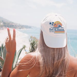May include: A white baseball cap with a "Gulf of Mexico" design, featuring palm trees and a sunset. The cap is worn by a person with long blonde hair, facing a beach with blue water and a clear sky. The person is giving a peace sign.