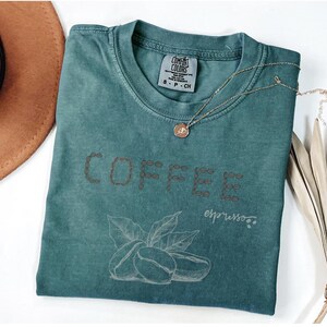 以下が含まれることがあります： 「COFFEE」の文字が茶色の糸で刺繍された、ティール色のComfort Colors Tシャツ。文字の下にはコーヒー豆と葉のグラフィックがあります。ペンダント付きのゴールドネックレスがシャツの上にあります。