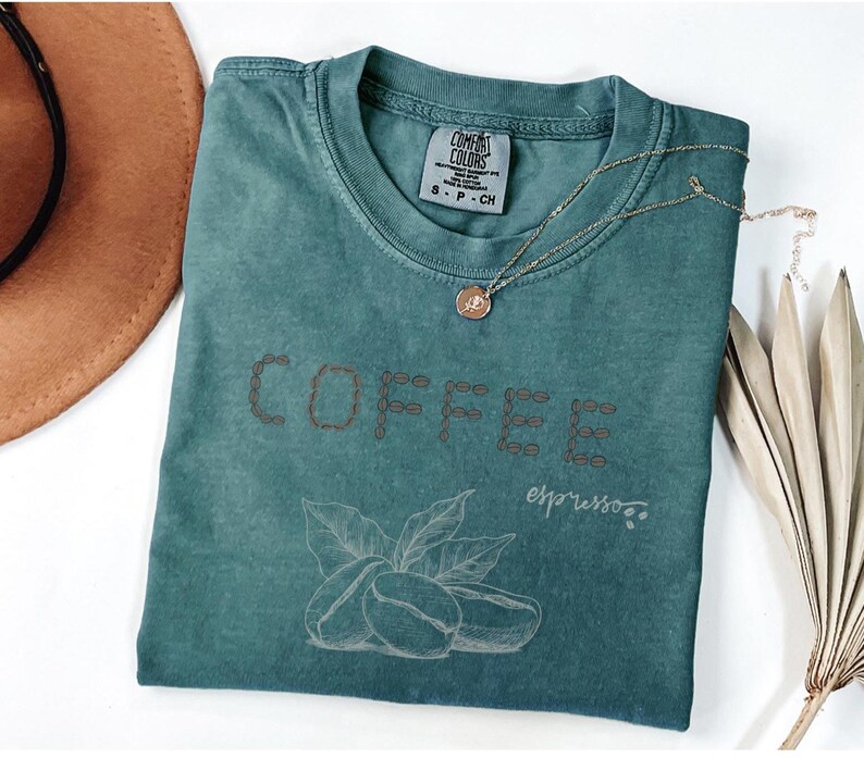 以下が含まれることがあります： 「COFFEE」の文字が茶色の糸で刺繍された、ティール色のComfort Colors Tシャツ。文字の下にはコーヒー豆と葉のグラフィックがあります。ペンダント付きのゴールドネックレスがシャツの上にあります。