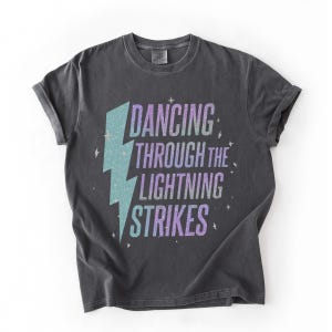 Pode incluir: Camiseta cinza escuro com mangas enroladas. A camiseta apresenta um raio turquesa e roxo e as palavras "DANCING THROUGH THE LIGHTNING STRIKES" em uma fonte estilizada. A camiseta tem um visual vintage e desgastado.