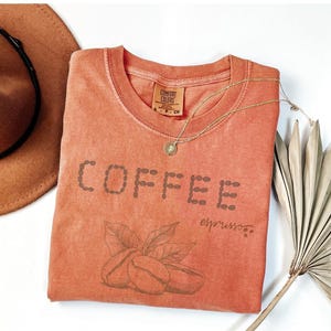 以下が含まれることがあります： コーヒー豆の形で「COFFEE」と書かれた、コーラル色のTシャツ。下にはコーヒー豆と葉があります。「espresso」の文字が下に書かれています。茶色の帽子と金のネックレスも画像にあります。