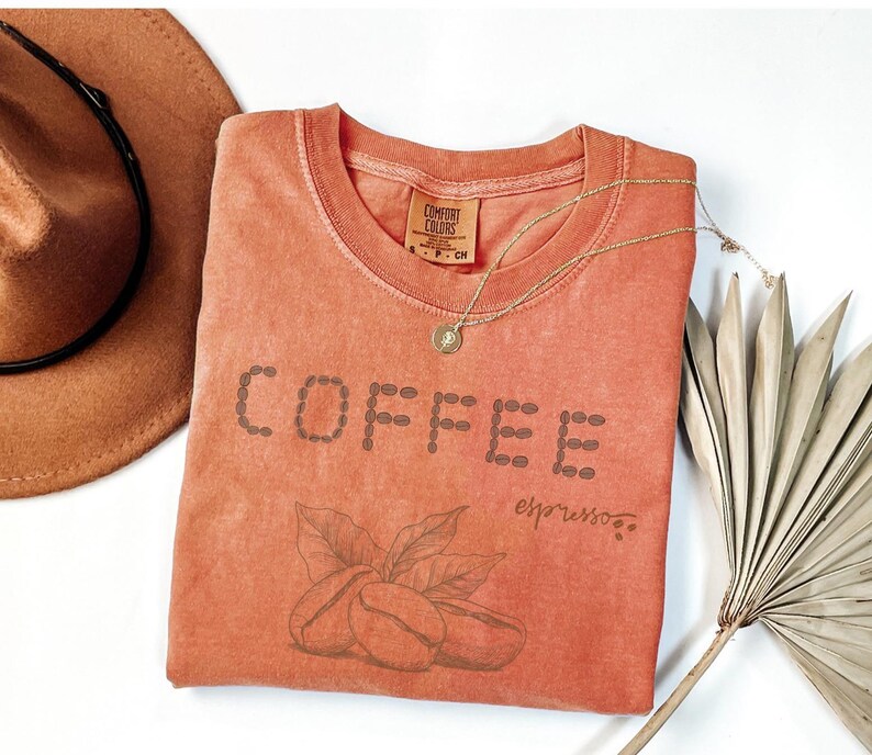 以下が含まれることがあります： コーヒー豆の形で「COFFEE」と書かれた、コーラル色のTシャツ。下にはコーヒー豆と葉があります。「espresso」の文字が下に書かれています。茶色の帽子と金のネックレスも画像にあります。