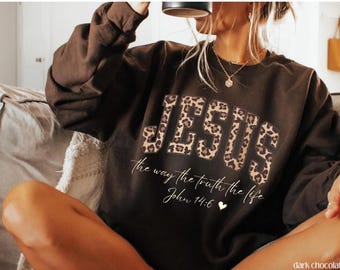 Op de afbeelding: Donkerchocoladekleurige sweatshirt met het woord "JESUS" in luipaardprintletters. Daaronder staat de zin "the way the truth the life" en de Bijbeltekst Johannes 14:6.