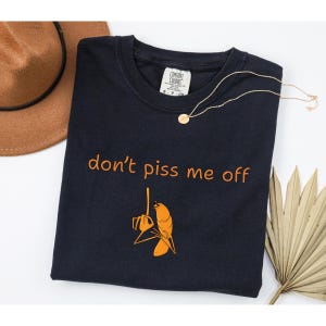 May include: A navy blue t-shirt with the text "don't piss me off" in orange, along with an orange graphic of a bird holding a chainsaw. A gold necklace and a brown hat are also visible.
