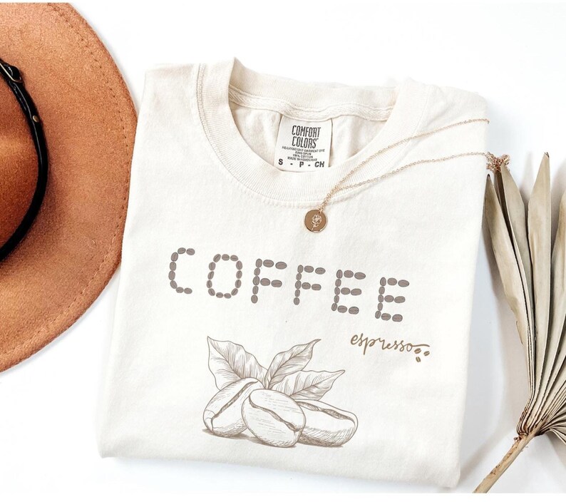 以下が含まれることがあります： クリーム色のTシャツ。点線で「COFFEE」の文字が描かれています。その下に葉っぱのついたコーヒー豆が2つ、筆記体で「espresso」の文字。金のネックレスと茶色の帽子も写っています。