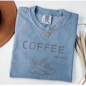 以下が含まれることがあります： コーヒー豆の形で「COFFEE」と書かれた水色のTシャツ。その下にコーヒー豆と葉のグラフィックがあります。金のネックレスがシャツの上に置かれています。