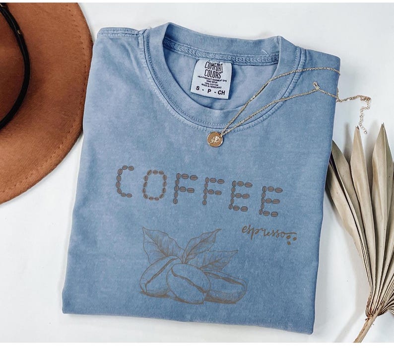 以下が含まれることがあります： コーヒー豆の形で「COFFEE」と書かれた水色のTシャツ。その下にコーヒー豆と葉のグラフィックがあります。金のネックレスがシャツの上に置かれています。