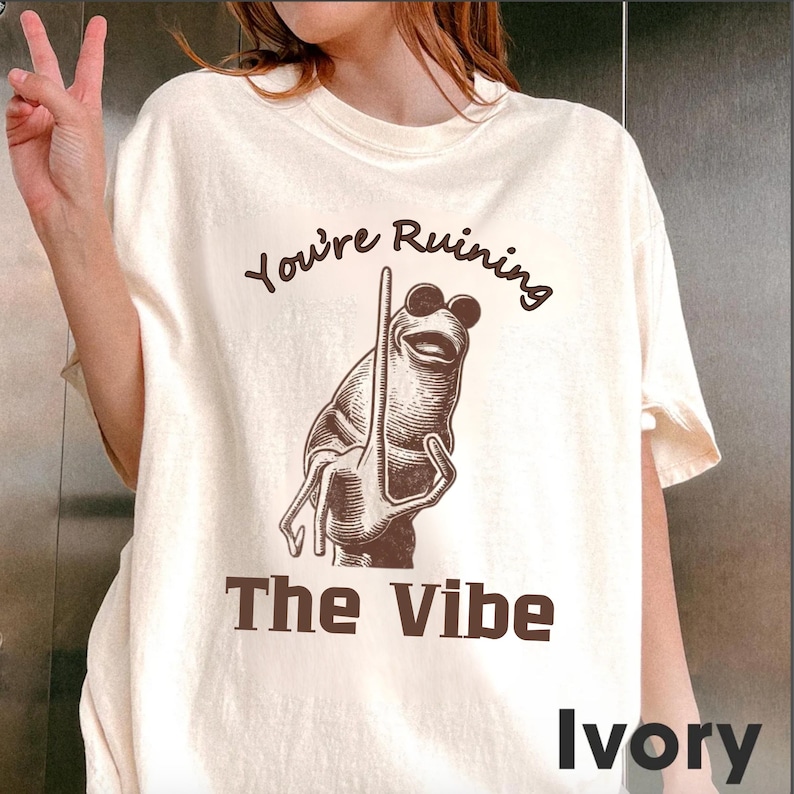 May include: Ivory-colored t-shirt with the text "You're Ruining The Vibe" and a cartoon frog illustration. The frog is brown and wearing sunglasses. The shirt is a casual, unisex style.