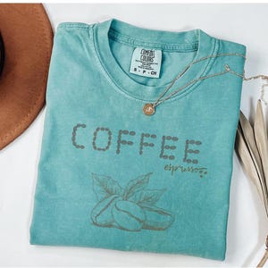以下が含まれることがあります： コーヒー豆の形で「COFFEE」と書かれた、ターコイズ色のComfort Colors Tシャツ。 「COFFEE」の下には筆記体で「espresso」と書かれています。 金色のペンダント付きネックレスがTシャツの上にあります。 コーヒー豆と葉のグラフィックが「COFFEE」の下にあります。