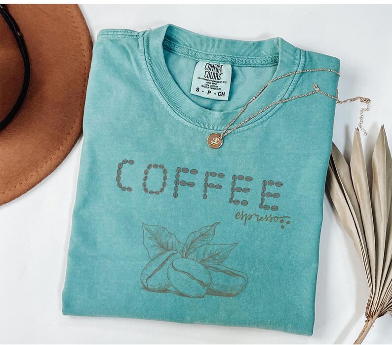 以下が含まれることがあります： コーヒー豆の形で「COFFEE」と書かれた、ターコイズ色のComfort Colors Tシャツ。 「COFFEE」の下には筆記体で「espresso」と書かれています。 金色のペンダント付きネックレスがTシャツの上にあります。 コーヒー豆と葉のグラフィックが「COFFEE」の下にあります。