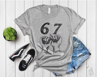 May include: A gray t-shirt with the number 67 in black, above two hands interlocked in a pinky promise. The shirt is paired with denim shorts and black sneakers on a white wooden surface.
