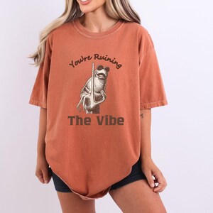 May include: Burnt orange t-shirt with the text "You're Ruining The Vibe" and a cartoon frog wearing sunglasses. The frog is holding a stick. The shirt has short sleeves and a relaxed fit.