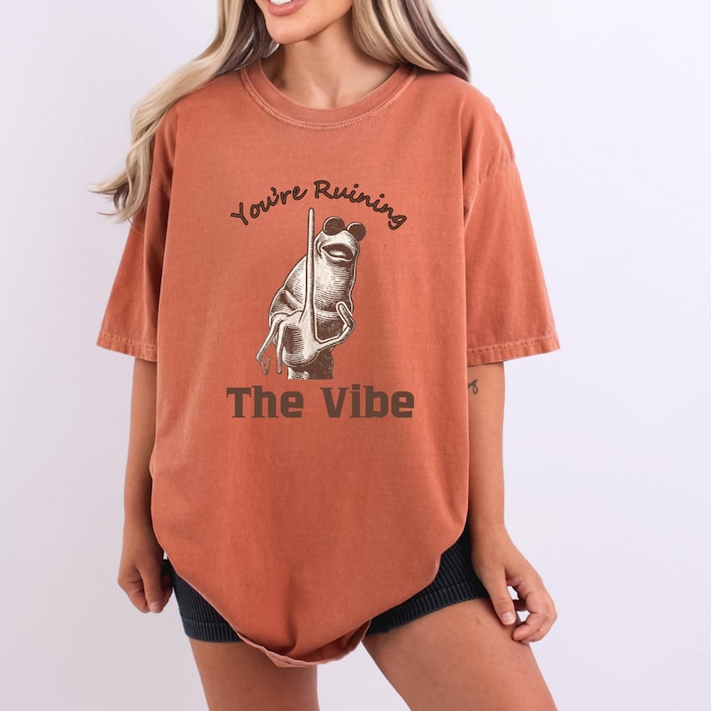 May include: Burnt orange t-shirt with the text "You're Ruining The Vibe" and a cartoon frog wearing sunglasses. The frog is holding a stick. The shirt has short sleeves and a relaxed fit.
