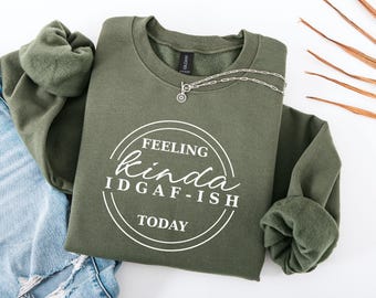 May include: An olive green sweatshirt with the text "FEELING kinda IDGAF-ISH TODAY" in a white circular design. A silver necklace with a pendant is draped across the sweatshirt. The sleeves are cuffed. The sweatshirt is placed on top of blue jeans.