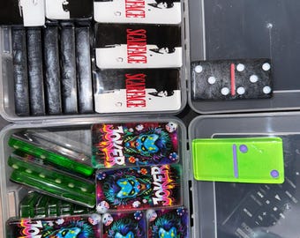 May include: A clear plastic container filled with custom dominoes. The dominoes feature designs inspired by the movie "Scarface" and the Joker. Some dominoes are green and translucent. A single black domino with white dots is also visible.
