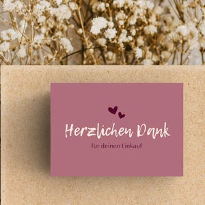 May include: A mauve-colored card with the words "Herzlichen Dank für deinen Einkauf" in white script, and two small purple hearts. The card is on a brown surface with dried white flowers in the background.