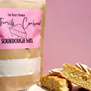 May include: A brown paper bag of "The Rosey Dough" Buttermilk Cornbread Sourdough Mix with a pink label. Sliced cornbread and whipped cream are on a plate. The background is a solid pink.