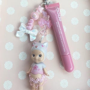 May include: A pink keychain featuring a small doll wearing a cat hat and a cherry blossom shirt, a tube of pink lip tint, and decorative pink and white charms. The lip tint tube reads "rhode PEPTIDE LIP TINT".