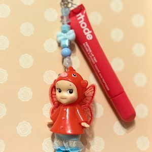 May include: A red and white keychain featuring a small doll in a red outfit with wings, a red lip tint tube, and a star-shaped charm. The keychain has blue and white beads.