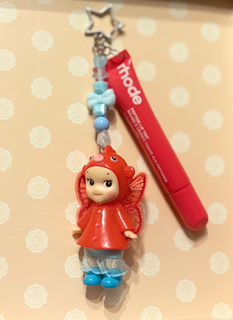 May include: A red and white keychain featuring a small doll in a red outfit with wings, a red lip tint tube, and a star-shaped charm. The keychain has blue and white beads.