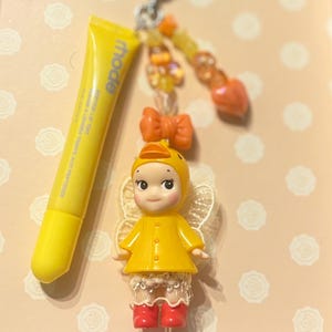 May include: A yellow lip tint tube and a charm featuring a doll in a yellow duck costume with red boots and lace trim. The charm has a beaded accent and a bow. The background is a light peach color.