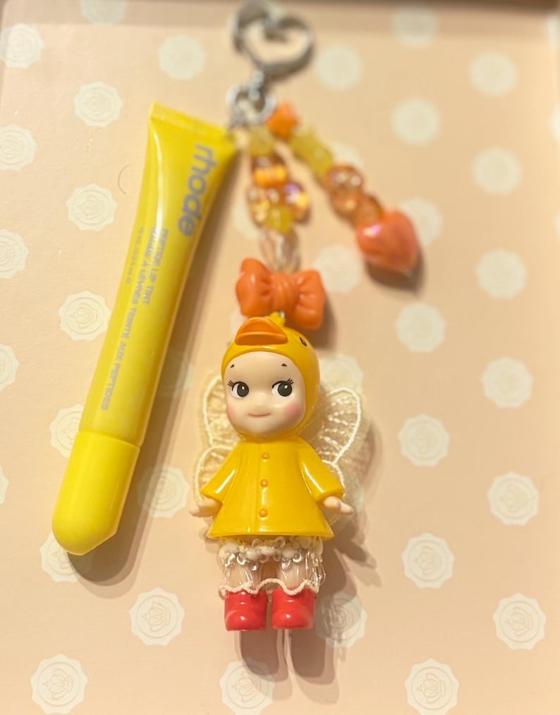 May include: A yellow lip tint tube and a charm featuring a doll in a yellow duck costume with red boots and lace trim. The charm has a beaded accent and a bow. The background is a light peach color.