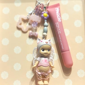 May include: A keychain with a small doll wearing a cat-eared hat and fairy wings, a pink lip tint tube, and decorative beads. The doll is wearing a pink shirt with a flower design. The lip tint tube has the word "rhode" printed on it.