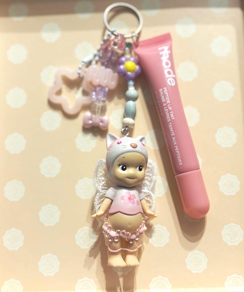 May include: A keychain with a small doll wearing a cat-eared hat and fairy wings, a pink lip tint tube, and decorative beads. The doll is wearing a pink shirt with a flower design. The lip tint tube has the word "rhode" printed on it.