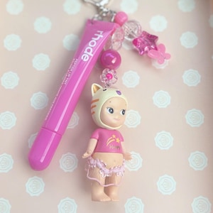 May include: A pink keychain featuring a lip tint tube with the brand name "rhode", a small doll wearing a cat-themed hat and a pink shirt with a star design, and decorative pink beads. The keychain is attached to a silver-colored clasp.