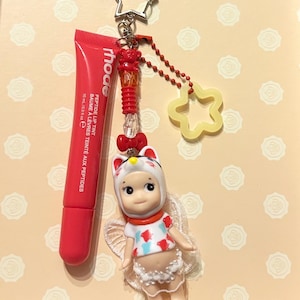 May include: A red lip tint tube and a charm featuring a doll in a cat costume, a star-shaped keychain, and a yellow star-shaped charm. The lip tint is labeled "rhode" and "PEPTIDE LIP TINT". The doll has white wings and a red bow.