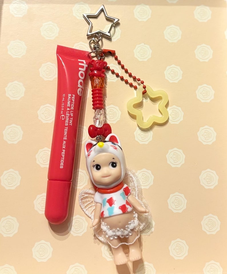 May include: A red lip tint tube and a charm featuring a doll in a cat costume, a star-shaped keychain, and a yellow star-shaped charm. The lip tint is labeled "rhode" and "PEPTIDE LIP TINT". The doll has white wings and a red bow.