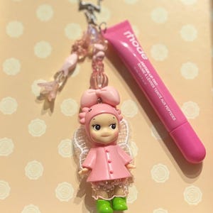 May include: A pink keychain featuring a doll with a bow, wings, and green boots, along with a tube of pink lip tint. The keychain has a star-shaped clasp and beaded accents. The lip tint tube reads "rhode".