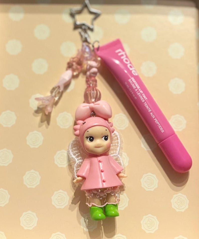 May include: A pink keychain featuring a doll with a bow, wings, and green boots, along with a tube of pink lip tint. The keychain has a star-shaped clasp and beaded accents. The lip tint tube reads "rhode".
