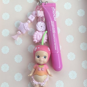 May include: A pink keychain with a heart-shaped clasp, adorned with pink beads and a small doll wearing a cherry-themed outfit. A tube of pink lip tint with the brand name "rhode" is attached. The lip tint tube is 10 ml / 0.3 oz.