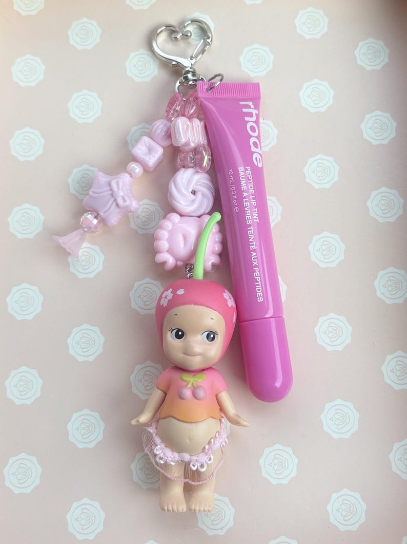 May include: A pink keychain with a heart-shaped clasp, adorned with pink beads and a small doll wearing a cherry-themed outfit. A tube of pink lip tint with the brand name "rhode" is attached. The lip tint tube is 10 ml / 0.3 oz.