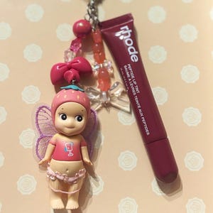 May include: A keychain featuring a small doll with a strawberry hat and fairy wings, a pink bow, and a tube of Rhode Peptide Lip Tint. The doll is wearing a pink shirt with a drink graphic. The keychain has a silver heart-shaped clasp.