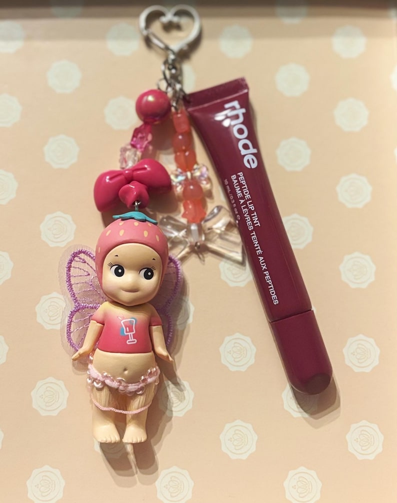 May include: A keychain featuring a small doll with a strawberry hat and fairy wings, a pink bow, and a tube of Rhode Peptide Lip Tint. The doll is wearing a pink shirt with a drink graphic. The keychain has a silver heart-shaped clasp.